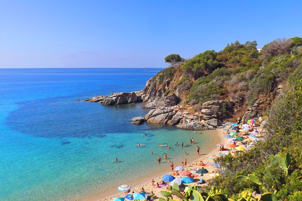 Turquoise cove beach with colourful umbrellas beneath green Mediterranean cliffs