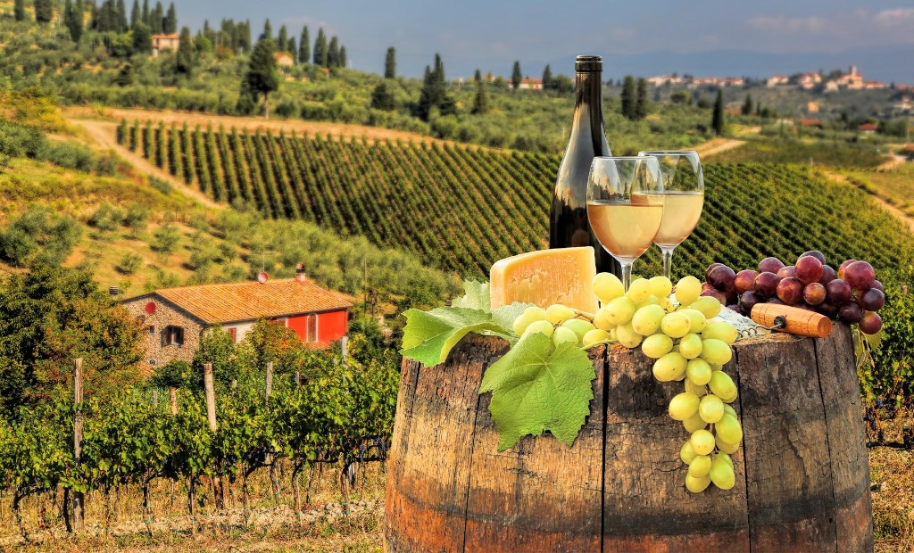 Wine barrel with glasses, cheese, and grapes overlooking vineyard hills and a stone farmhouse